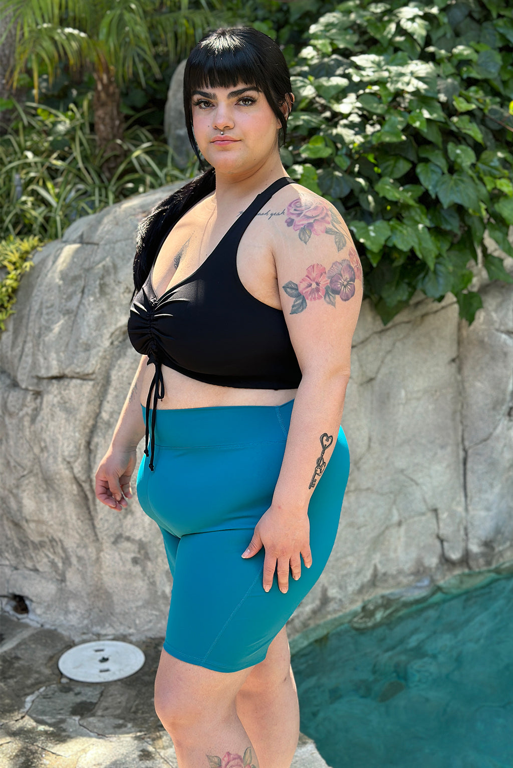 Black-haired model poses by pool and ivy wearing plus size 9 inch swim shorts in teal color