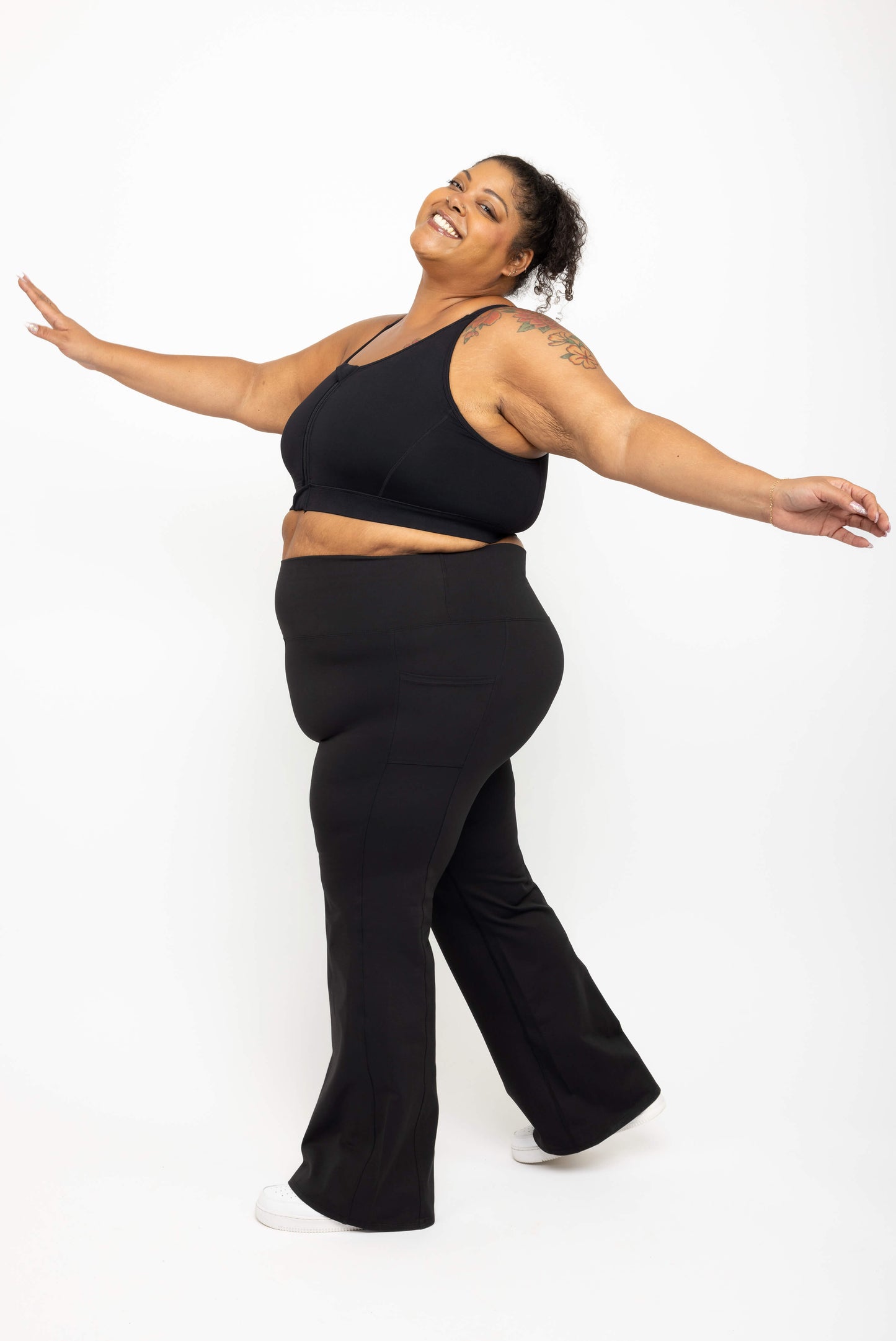 Profile view of model smiling and wearing Superfit Hero black plus size flare leggings with pockets size 3X