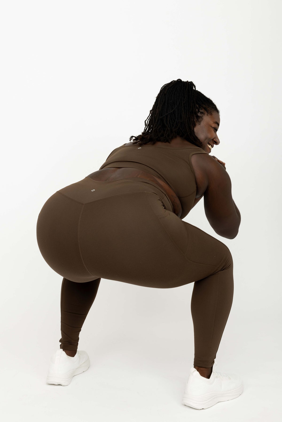 A woman wearing Chocolate Brown SuperHold Ankle Leggings, posing in a deep squat on a seamless white studio background.