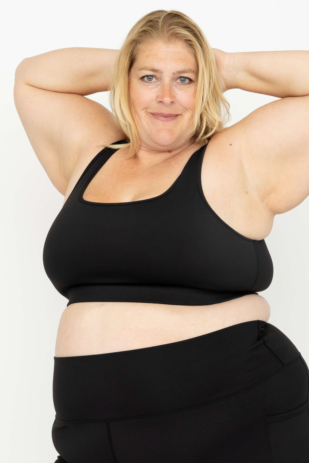 A plus-size woman wearing Superfit Hero SuperHold Black Scoop Bra, posing with her hands behind her head against a plain white background.