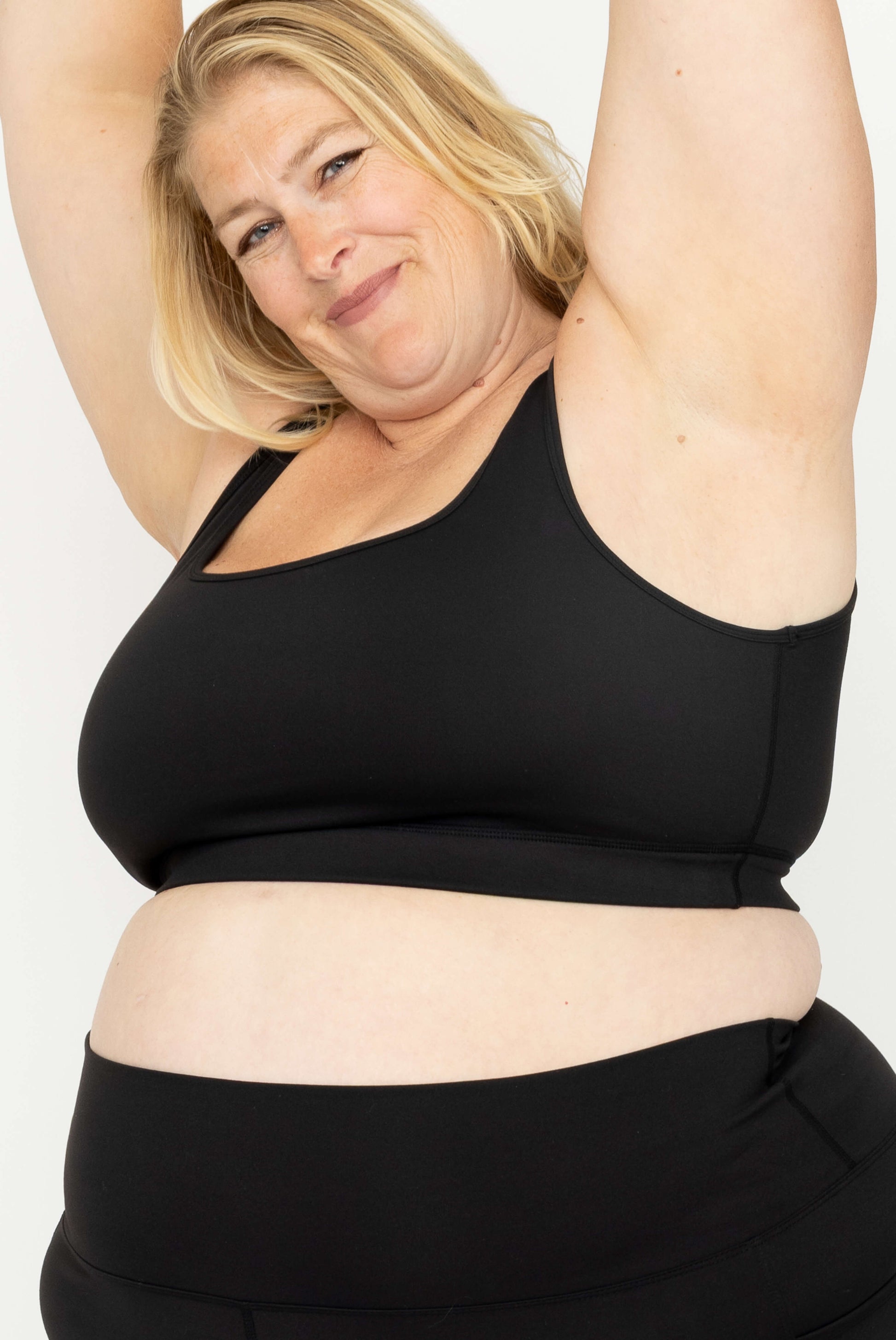 A woman wearing a Superfit Hero SuperHold Black Scoop Bra and matching black bottoms, posing with her arms raised to show full coverage under the arm. 