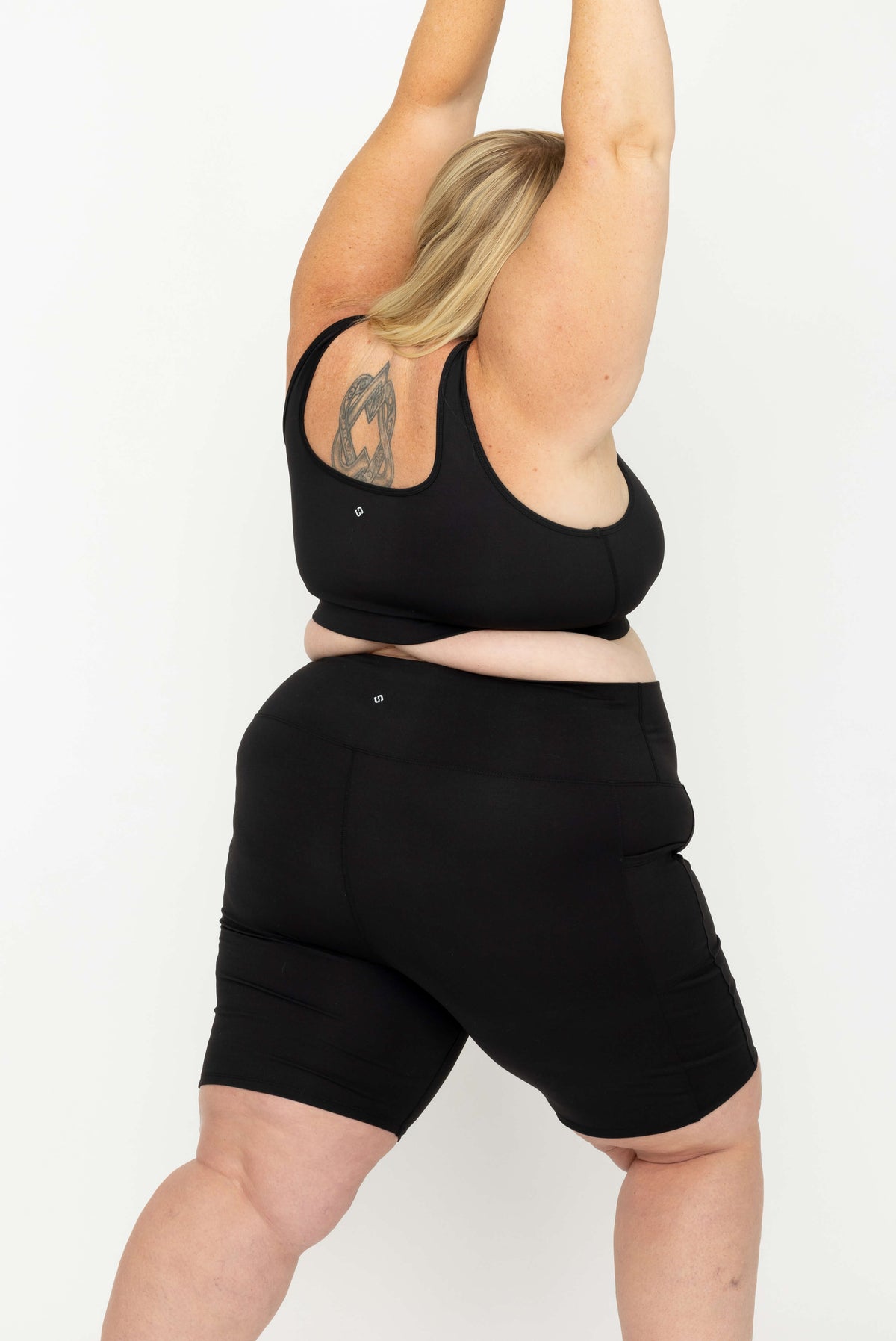 A plus-size woman wearing a Superfit Hero SuperHold Black Scoop Bra and matching black bike shorts, shown from the back against a plain white studio background.