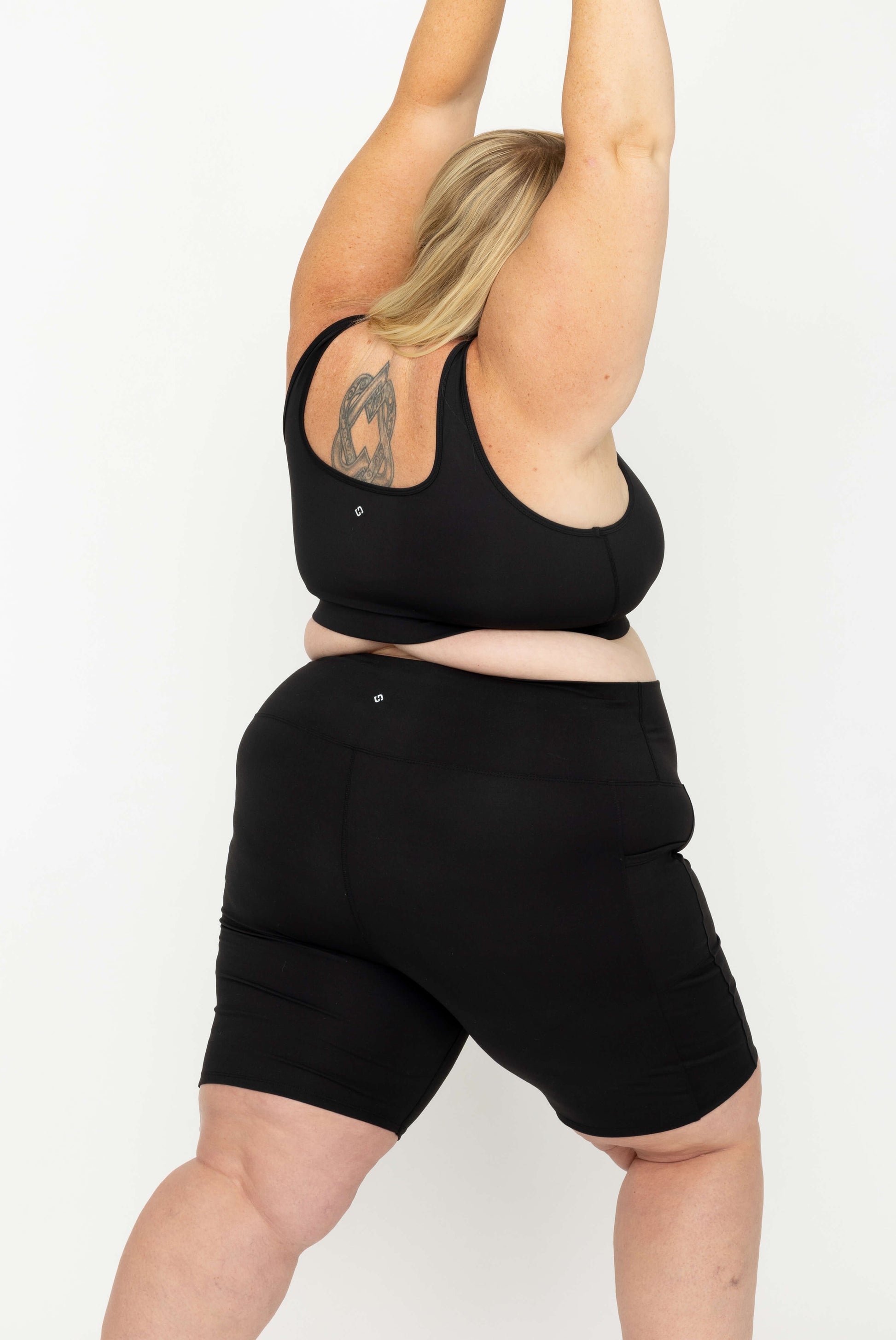 A plus-size woman wearing a Superfit Hero SuperHold Black Scoop Bra and matching black bike shorts, shown from the back against a plain white studio background.