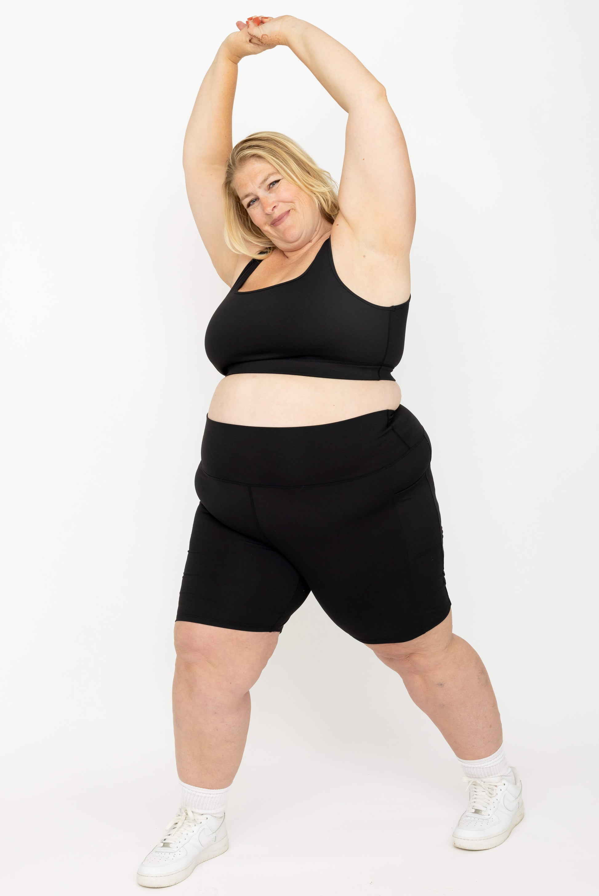 A plus-size woman wearing a Superfit Hero SuperHold Black Scoop Bra and matching black bike shorts, posing against a plain white studio background.