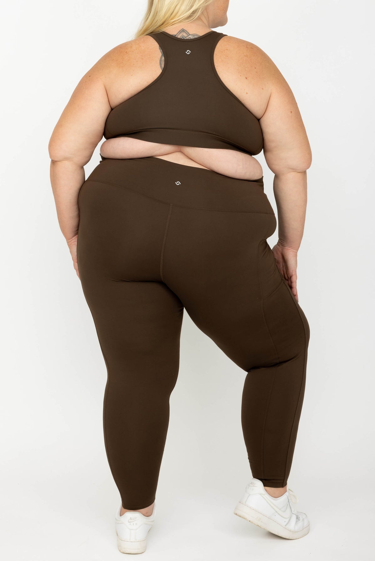 A plus-size model shown from the back wearing a matching activewear set, consisting of a racerback sports bra and high-waisted Chocolate Brown SuperHold 7/8 leggings, paired with white sneakers against a plain white studio background.