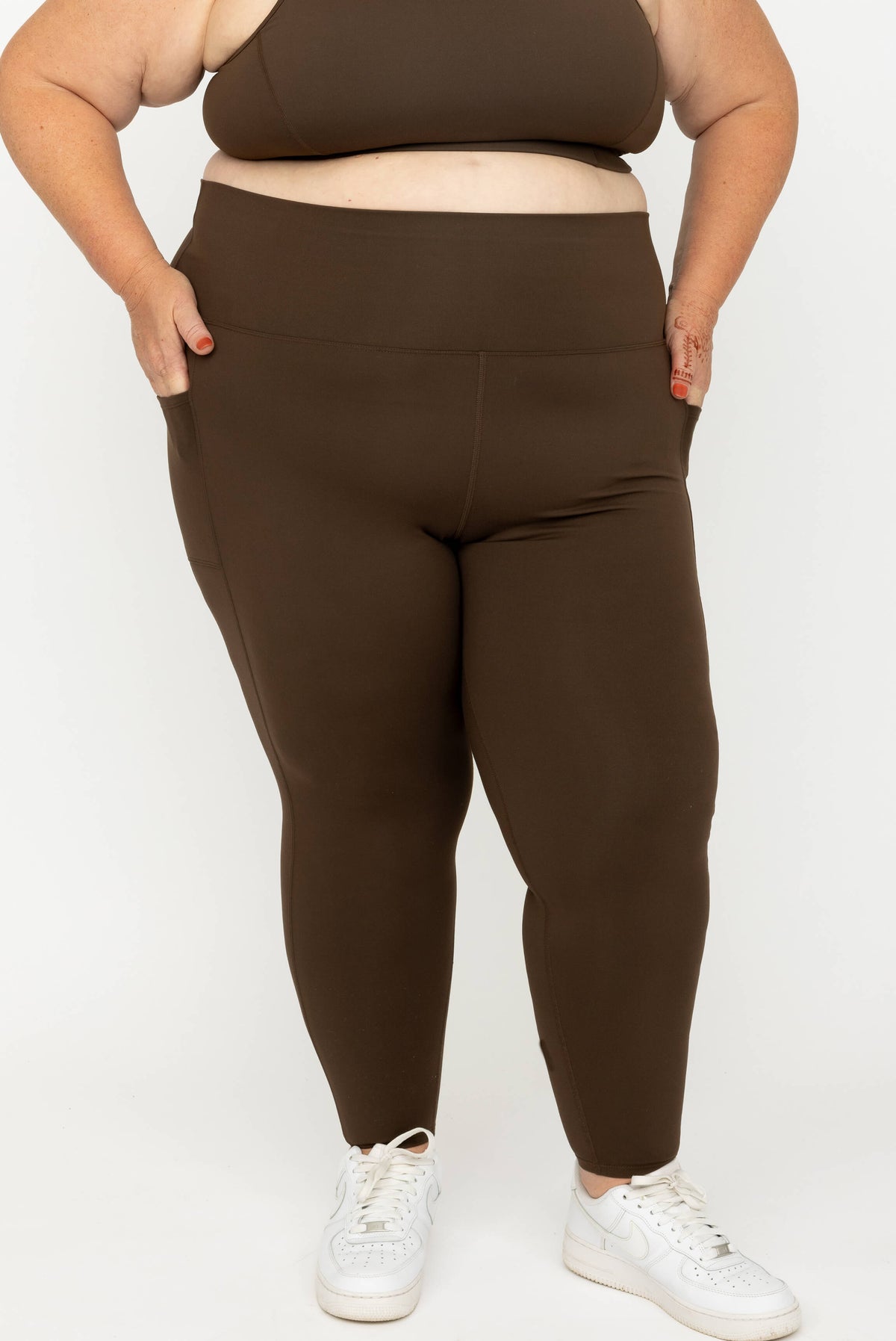 A plus-size model wearing high-waisted, Chocolate Brown SuperHold 7/8 leggings with side pockets, paired with a matching brown sports bra and white sneakers, against a plain white studio background.