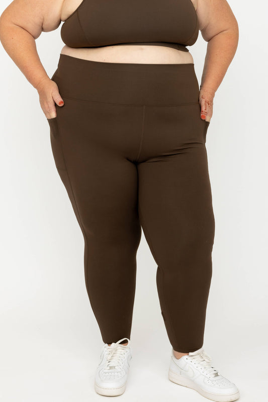 A plus-size model wearing high-waisted, Chocolate Brown SuperHold 7/8 leggings with side pockets, paired with a matching brown sports bra and white sneakers, against a plain white studio background.