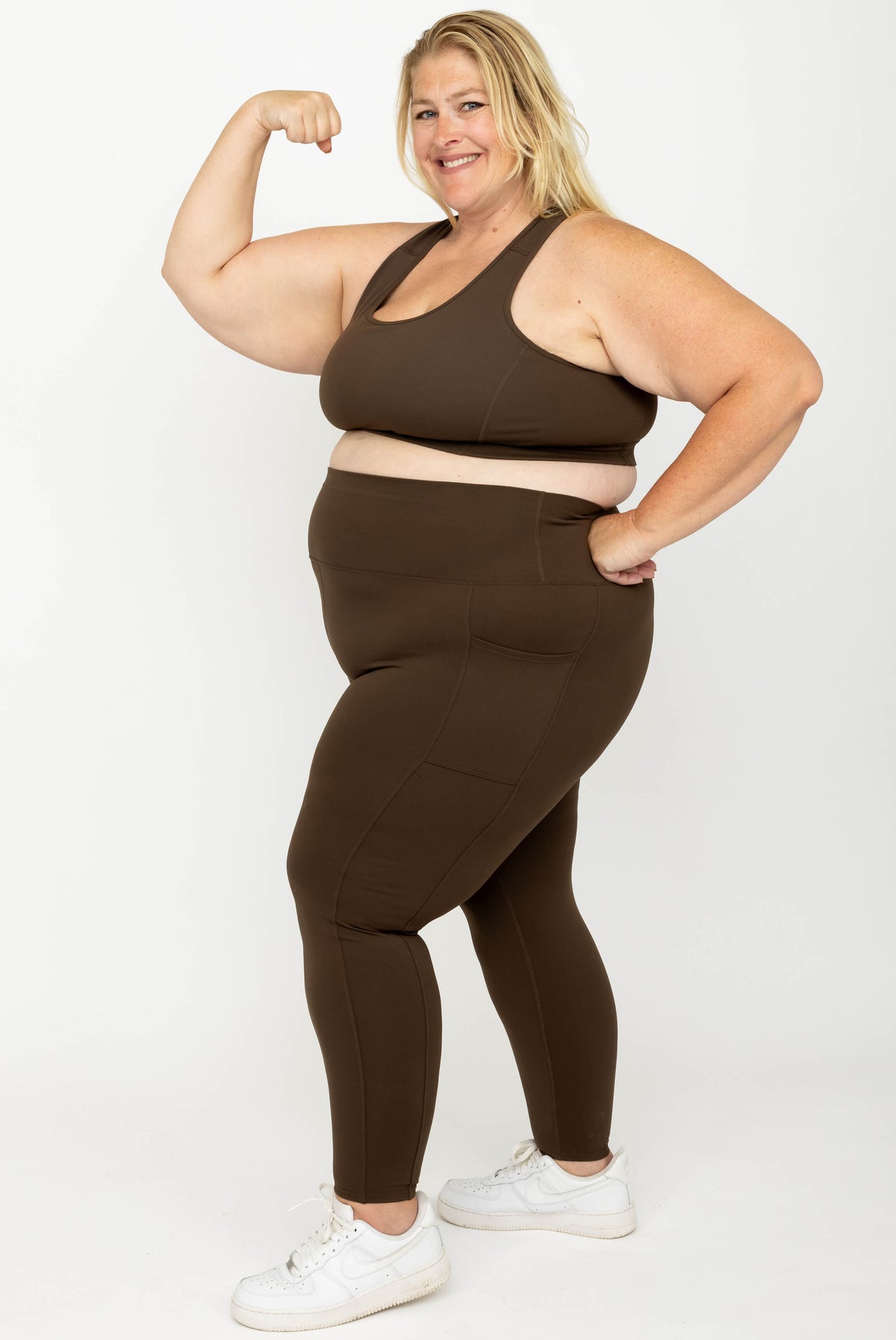 A woman modeling a matching activewear set consisting of a sports bra and high-waisted Chocolate Brown SuperHold 7/8 leggings, paired with white sneakers, against a plain white studio background.