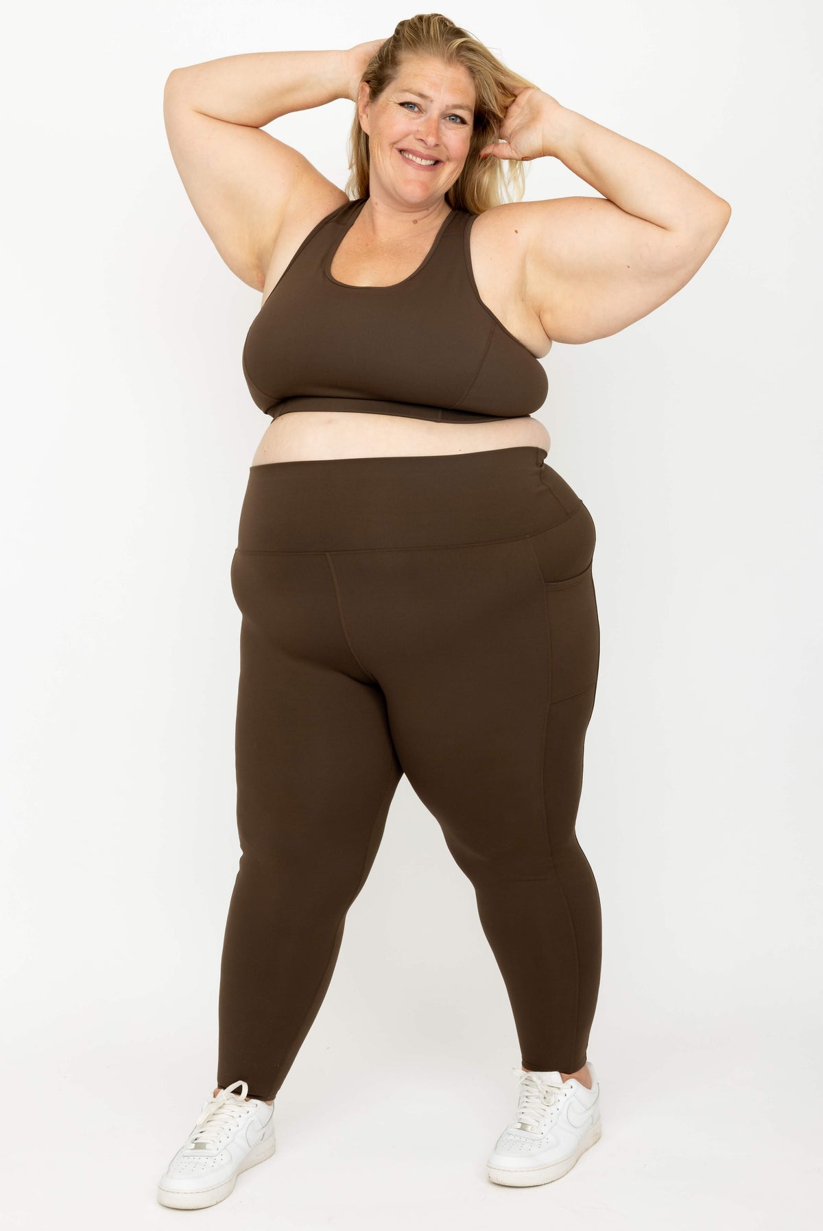 A plus-size woman modeling a matching dark brown activewear set consisting of SuperHold Racerback Sports Bra and high-waisted leggings, paired with white sneakers, against a plain white studio background.