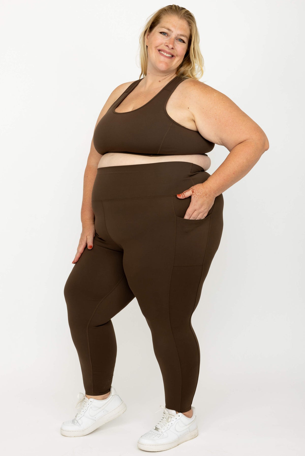 A plus-size woman modeling a matching  activewear set consisting of a sports bra and high-waisted Chocolate Brown SuperHold 7/8 leggings with side pockets, paired with white sneakers, against a plain white studio background.