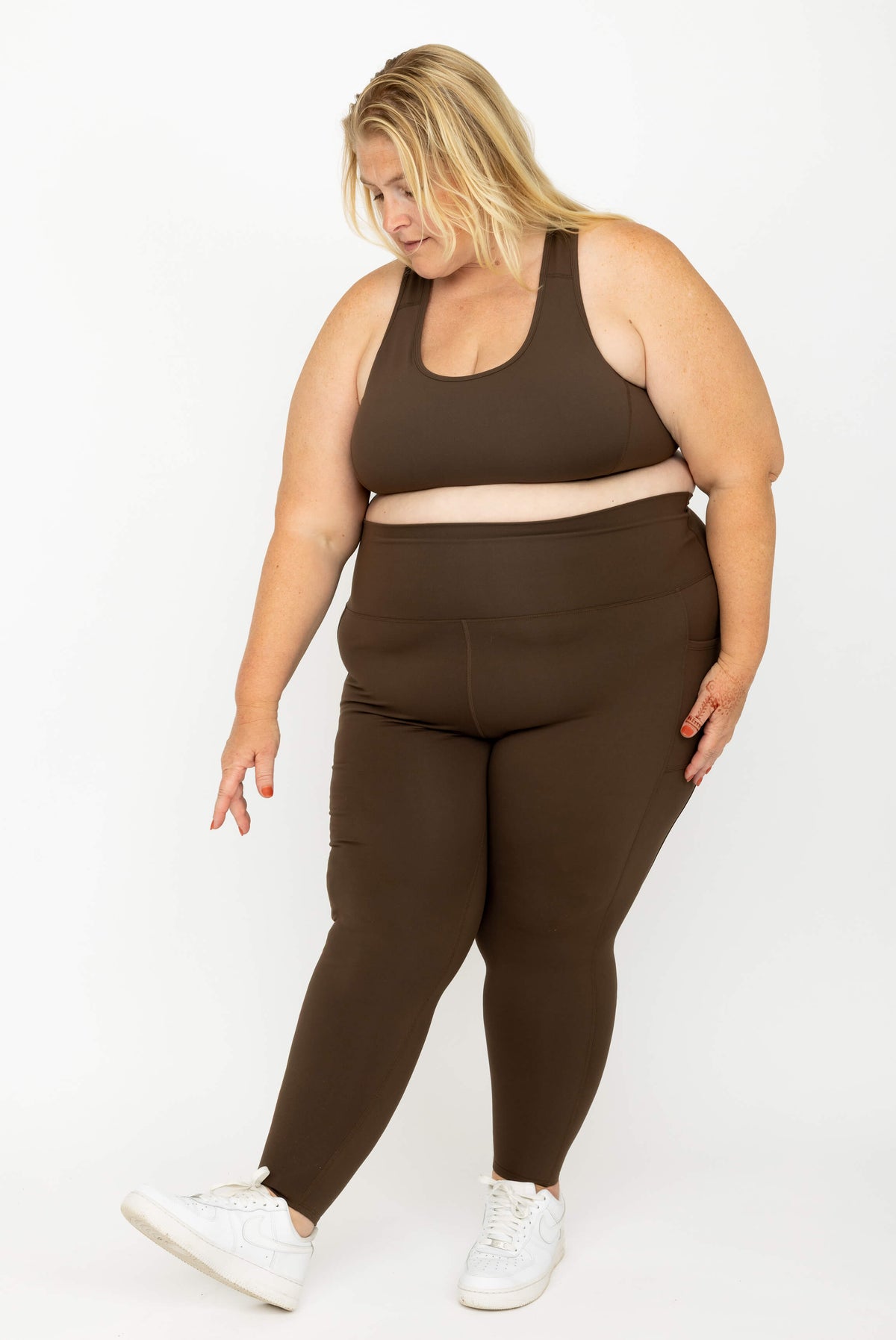 A plus-size woman modeling a matching dark brown activewear set consisting of a sports bra and high-waisted SuperHold 7/8 leggings, paired with white sneakers, against a plain white studio background.