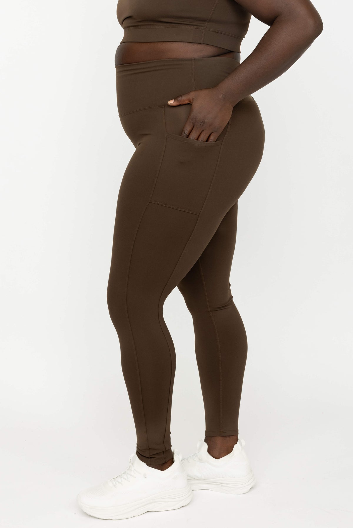 Side-view of model wearing Chocolate Brown SuperHold Ankle Legging with pockets, paired with white sneakers against a plain white studio background.
