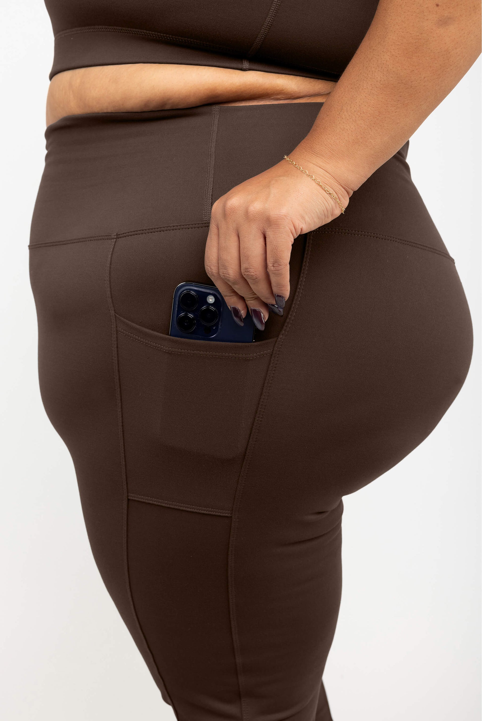 Close-up of a model wearing Chocolate brown high-waisted leggings with a side pocket holding a blue smartphone, against a plain light background.