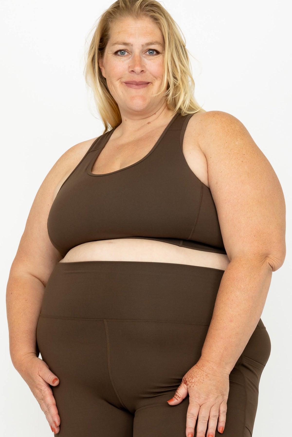 A plus-size blonde woman modeling a matching dark brown activewear set consisting of a SuperHold Racerback Sports Bra and high-waisted leggings, set against a plain white studio background.