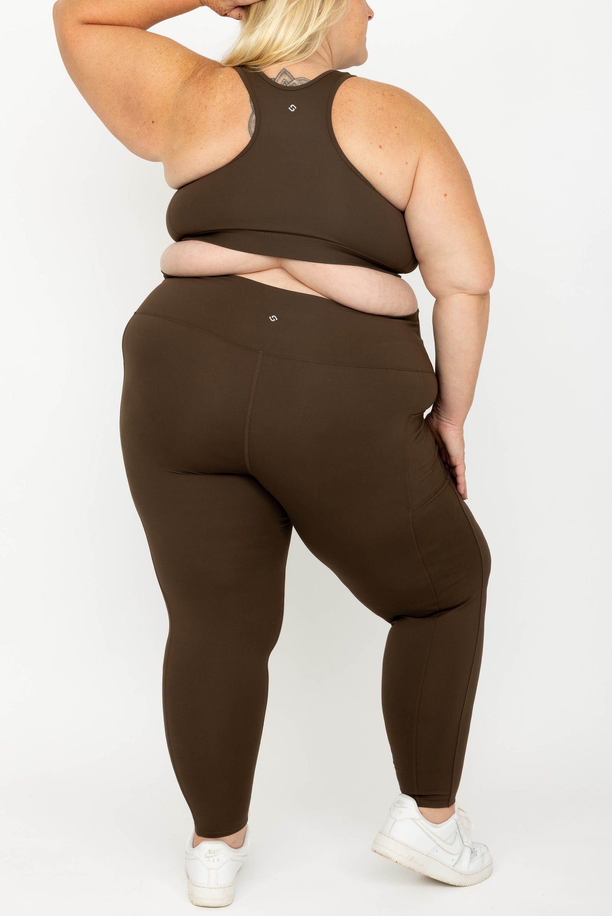 A plus-size model shown from the back wearing a matching dark brown activewear set consisting of a SuperHold Racerback Sports Bra and high-waisted leggings, paired with white sneakers against a plain white studio background.