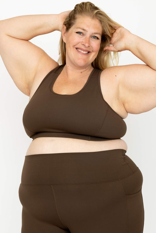 A smiling woman with blonde hair poses in a matching Superfit Hero dark brown activewear set, featuring SuperHold Racerback Sports Bra and high-waisted leggings, against a plain white background.