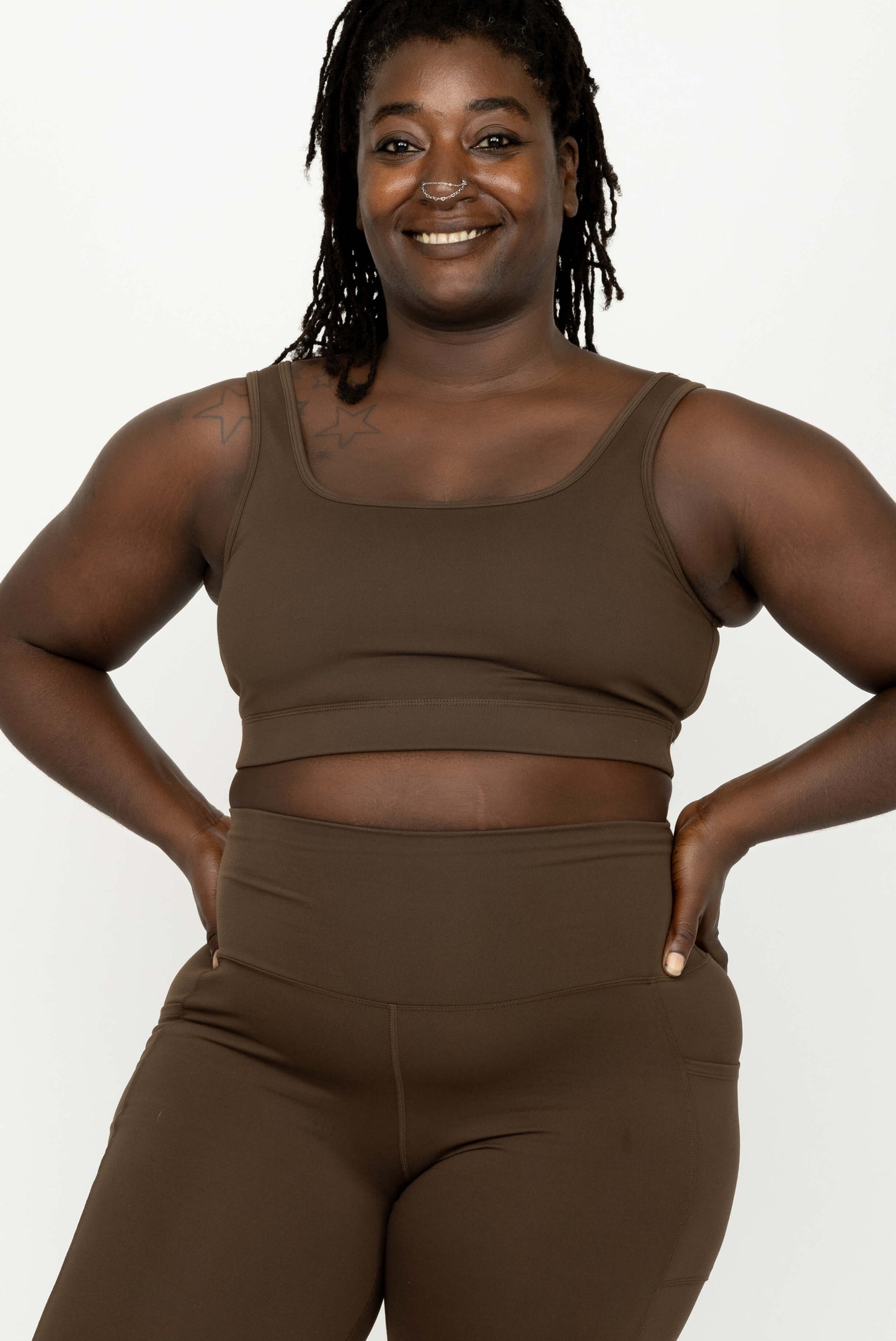 A smiling woman modeling a matching brown activewear set consisting of a Superfit Hero SuperHold Scoop Bra and high-waisted leggings, set against a plain white studio background.