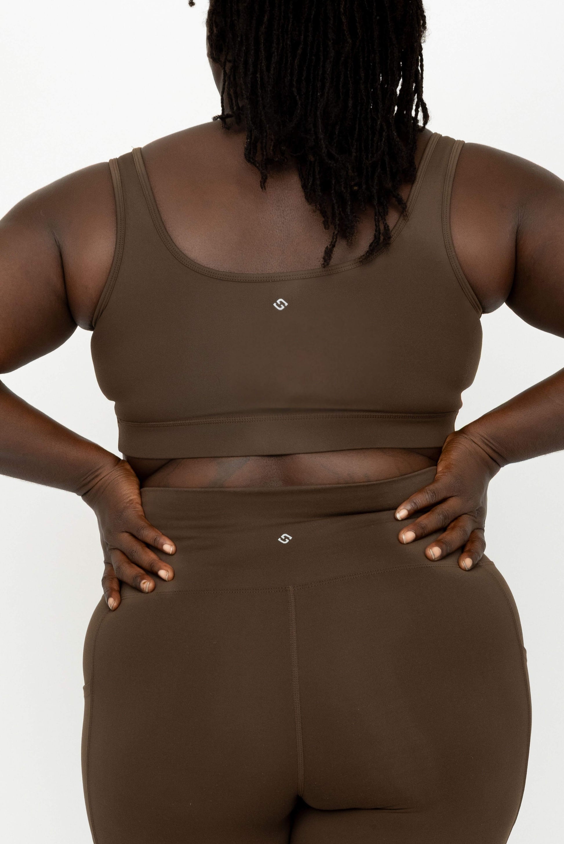 A close-up back view of a model wearing a matching brown activewear set, featuring a SuperHold Scoop Bra and high-waisted leggings with a small Superfit Hero logo at the center of each garment.