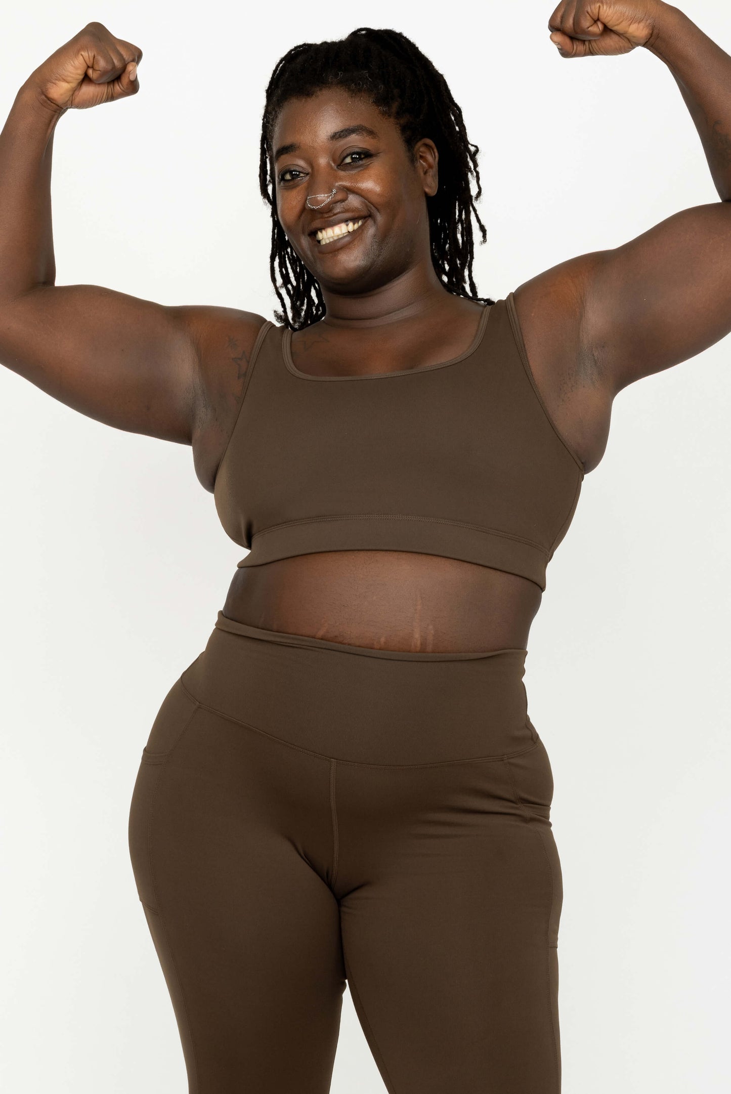 A smiling woman with braided hair poses in a Superfit Hero Chocolate Brown SuperHold Scoop Bra and high-waisted leggings set against a plain light background.