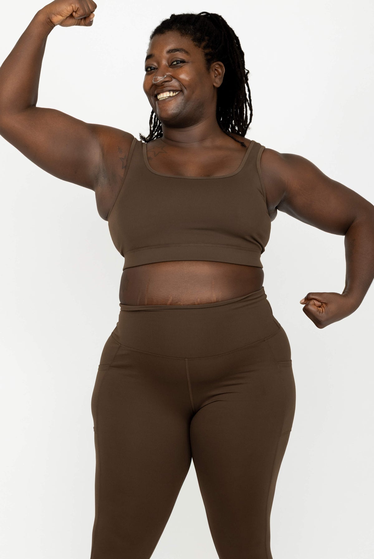 A smiling woman modeling a matching brown activewear set consisting of Superfit Hero SuperHold Scoop Bra and high-waisted leggings, posing against a white background.