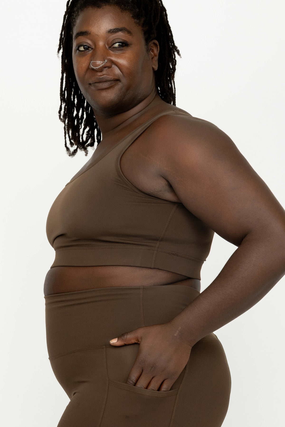 A plus-size woman wearing a matching brown activewear set consisting of a Superfit Hero SuperHold Scoop Bra and high-waisted leggings, shown from a side profile against a plain white background.