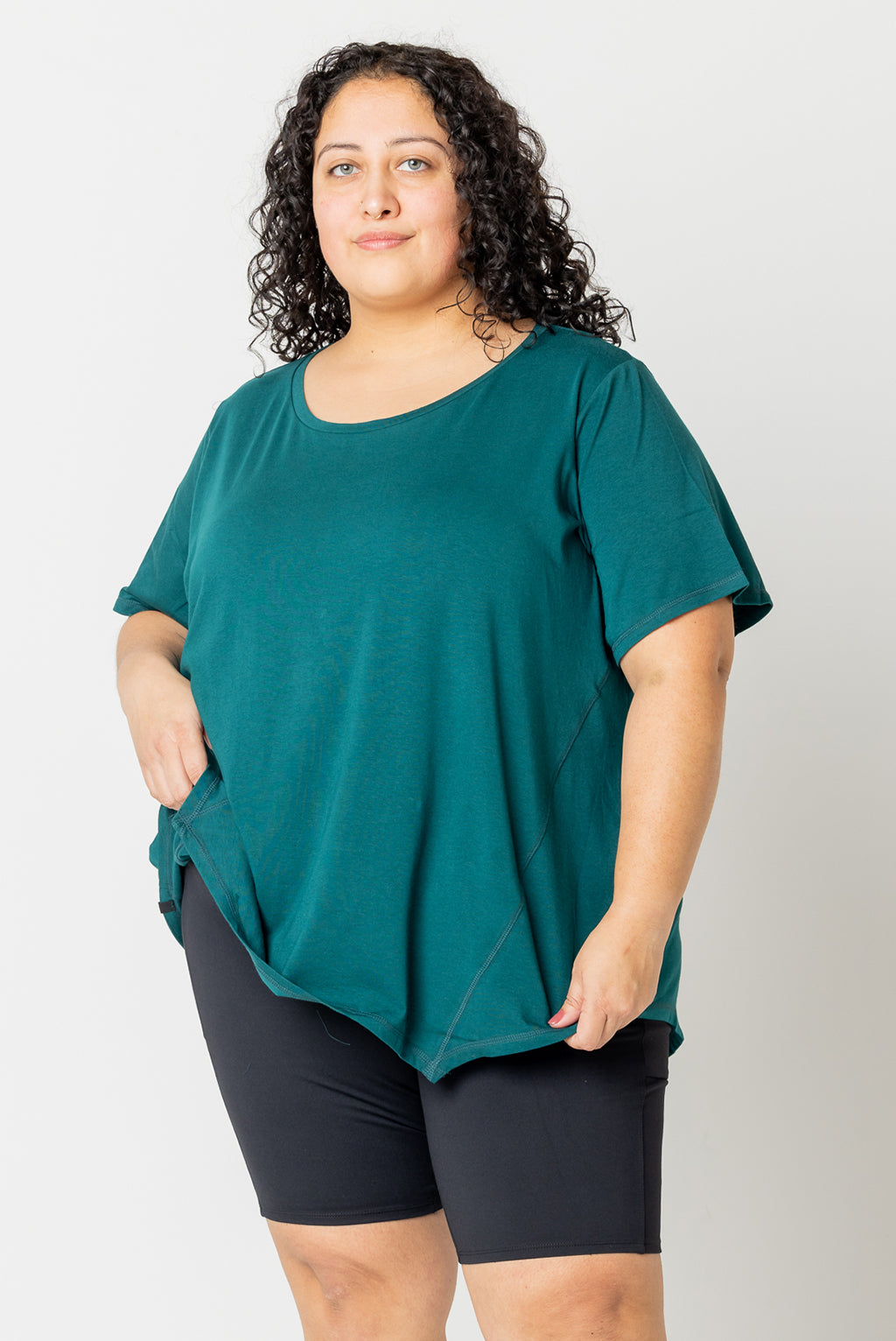 Front view of model holding up one side of hem of plus size flow tee in forest green size 3X