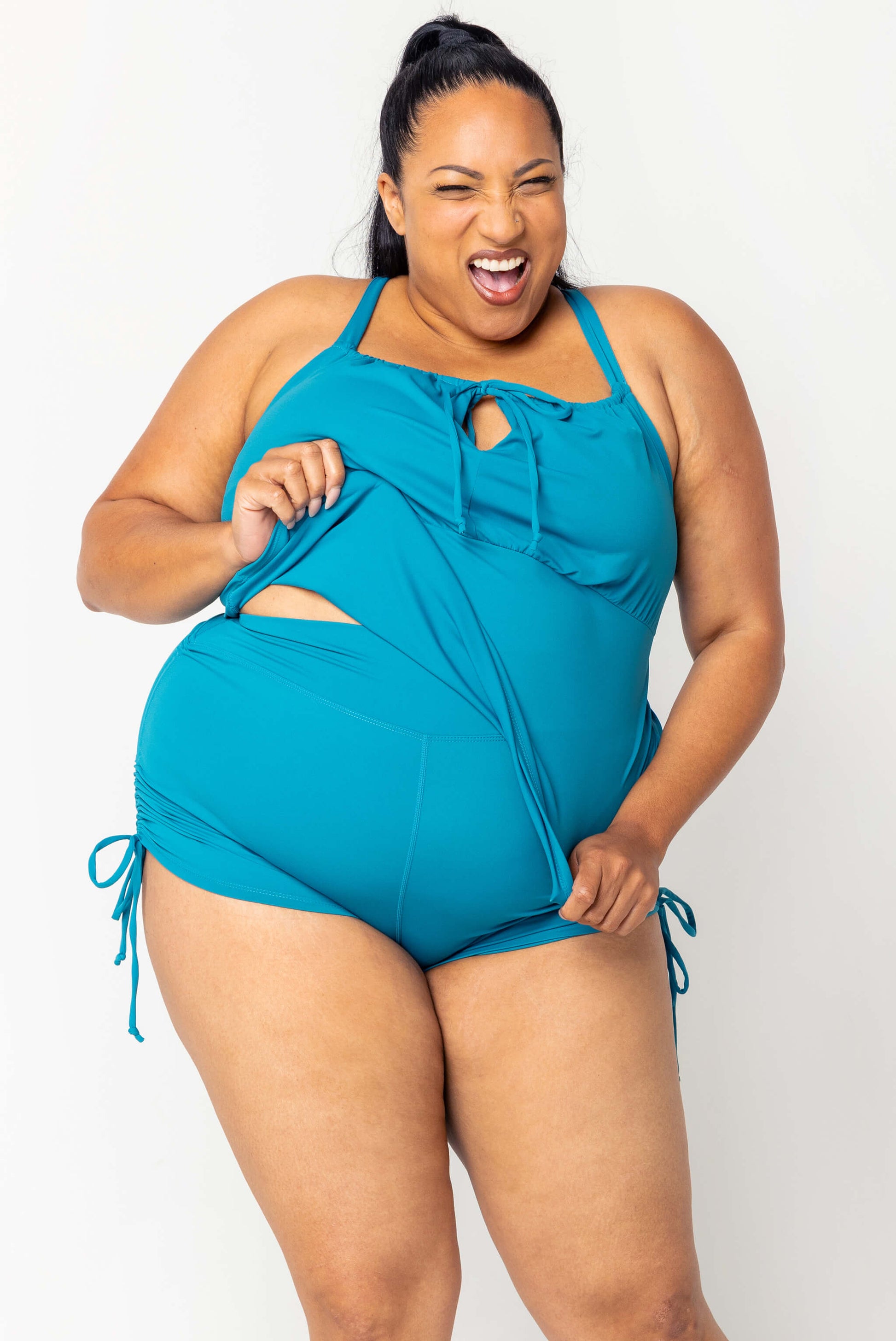 Model pulling the hem of teal swim tankini to show the matching booty shorts underneath