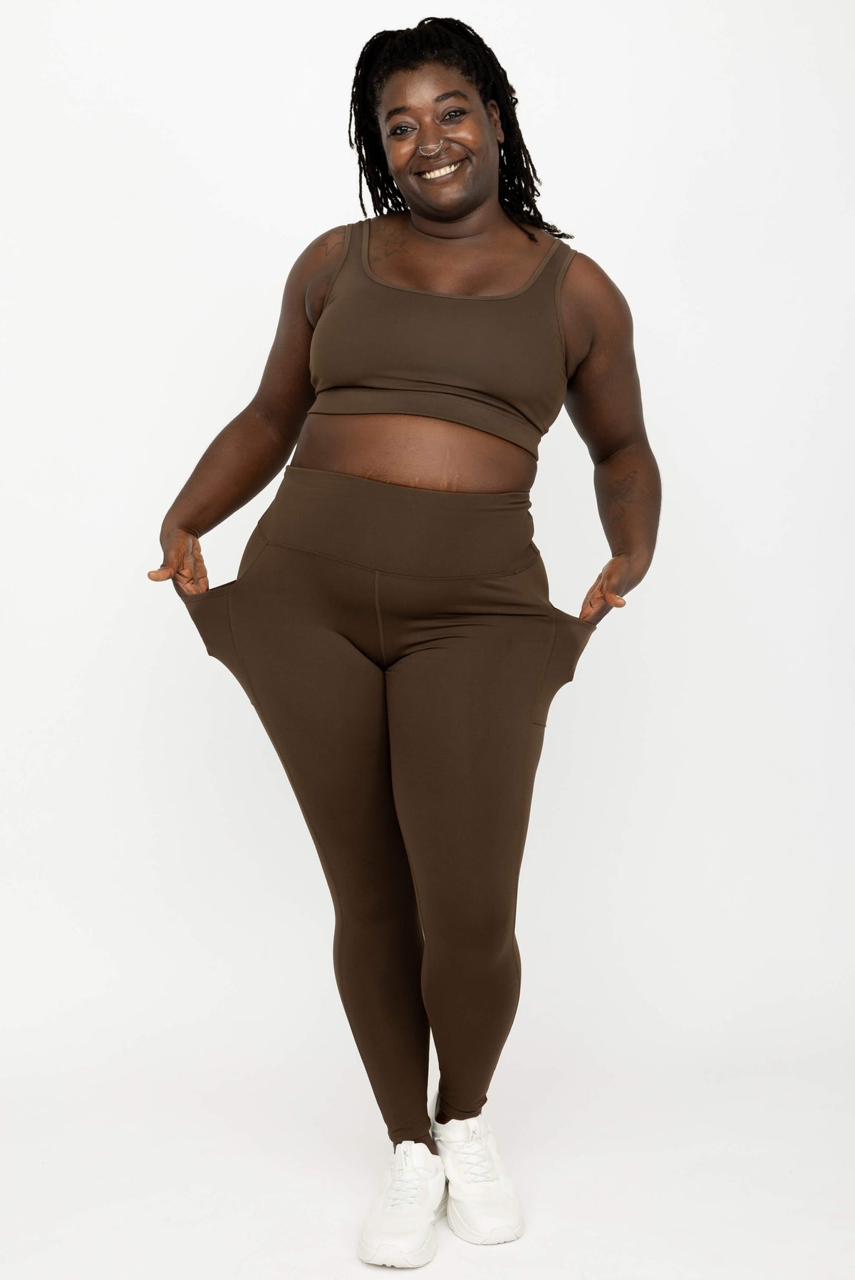 A model wearing a matching activewear set consisting of a sports bra and high-waisted Chocolate Brown SuperHold Ankle Leggings, posing with her hands in her pockets.