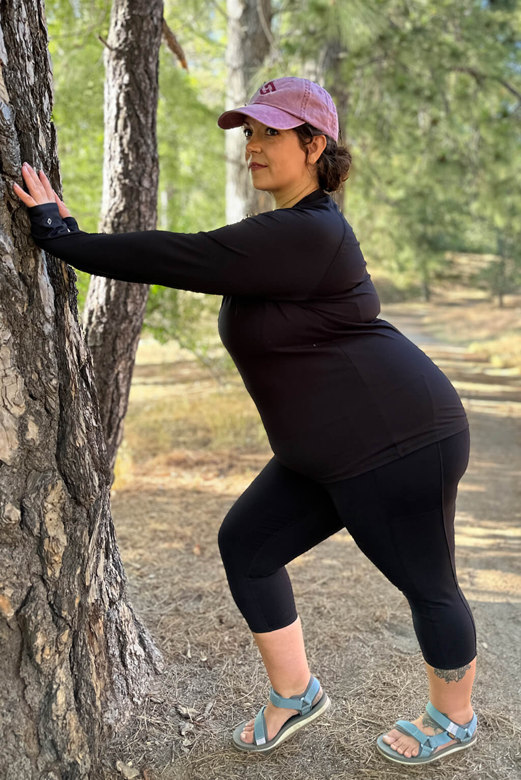 Plus size model stretches against a tree wearing black long sleeve compression top.
