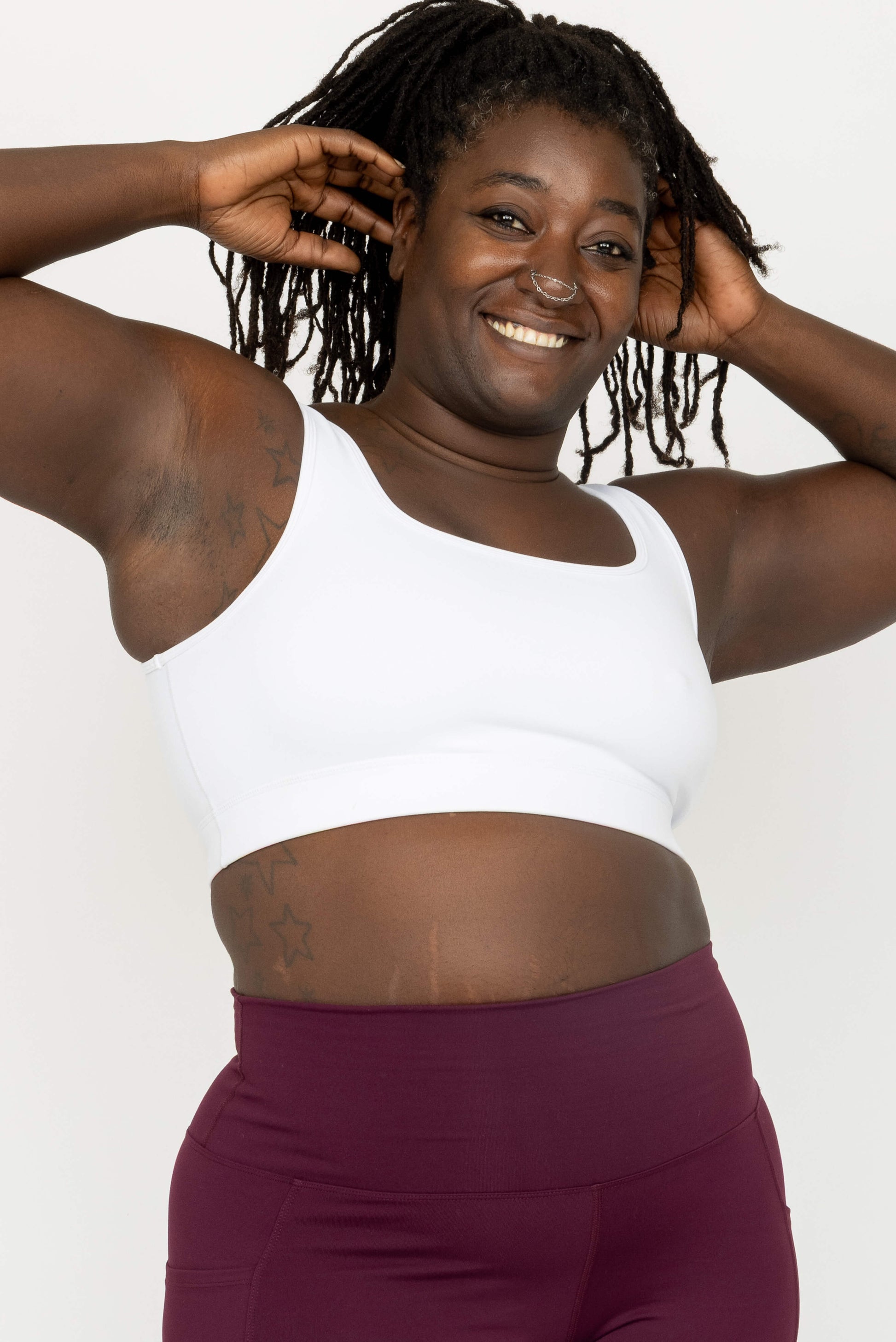 A smiling woman with dark braids poses in a White SuperHold Scoop Bra and burgundy leggings against a clean white studio background.