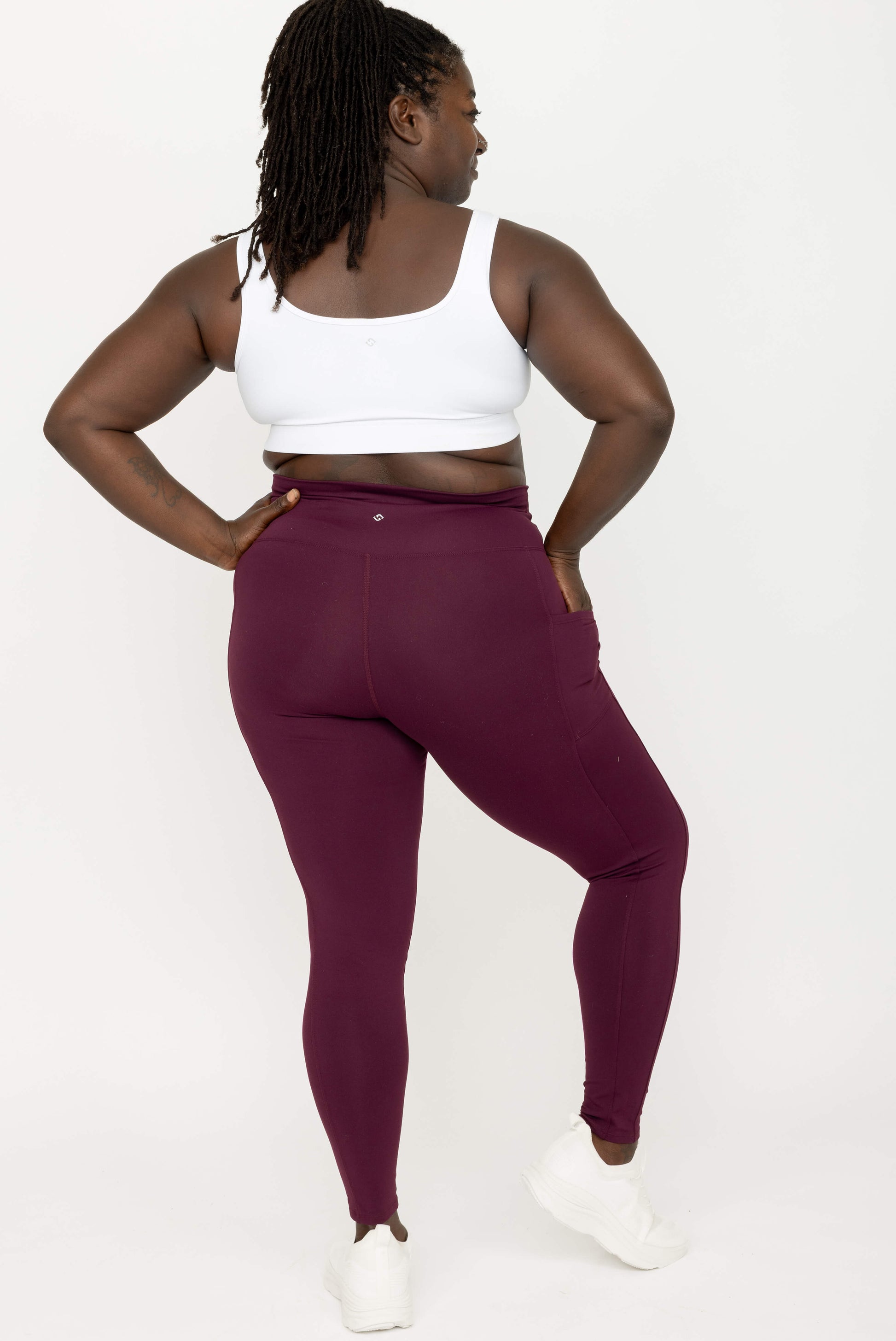 A woman modeling high-waisted leggings in a deep burgundy color, paired with a white sports bra and white sneakers, shown from a back-three-quarter angle against a plain white studio background.